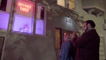 a couple stands in front of a window with a sign in it that says "distance tarot" and a woman below the sign wearing a pointy wizard's hat.