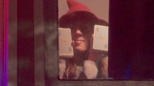 A woman wearing a hat holds up two piles of cards in her two hands from behind a window