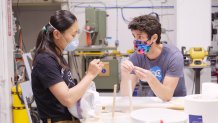 a man and a woman stand at a workbench talking, with masks covering their lower faces