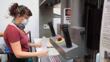 a woman stands at a large machine, holding two five-sided pieces of clear plastic in her hands