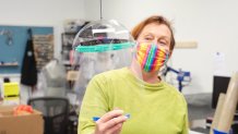 a woman with a colorful mask over her nose and mouth holds up a clear plastic face shield