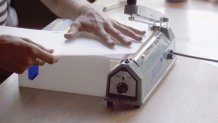 a woman's hands align a five-sided piece of clear plastic with the hot edge of a heat sealing machine