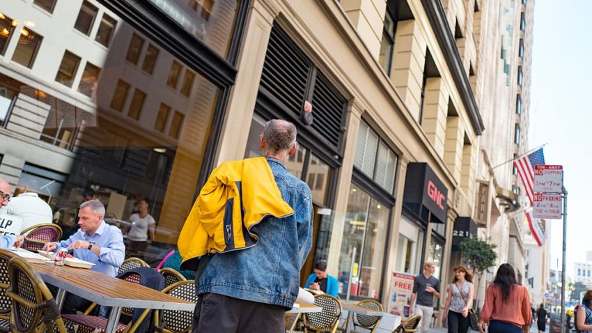View of a homeless man, in a denim jacket, near a sidewalk restaurant where a man in a button down shirt eats lunch, in the Financial District neighborhood of San Francisco, California, September 26, 2016. (Photo via Smith Collection/Gado/Getty Images).