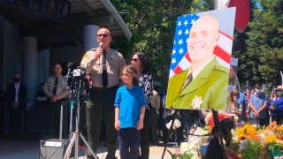 Santa Cruz Sheriff Jim Hart stands next to a photo of fallen Sgt. Damon Gutzwiller, next to the officer's pregnant widow and child as more than a thousand people gather outside the Santa Cruz County Sheriff-Coroner's Office to pay their respects in Santa Cruz, Calif., Sunday, June 7, 2020.