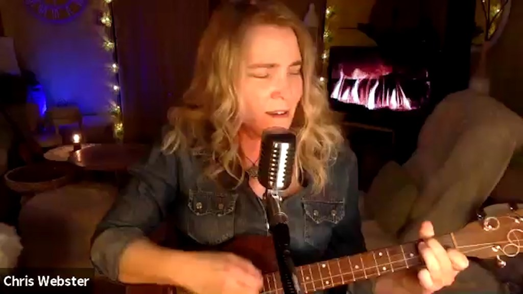 A woman plays a ukulele in front of a digital fireplace with christmas lights in the background