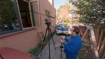 Reporter wearing a facemask stands outside a window with a tripod and microphone stand, interviewing a woman sitting in her home.