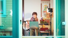 A woman sits at a laptop with her guitar. The camera is looking into her home from outside a window.