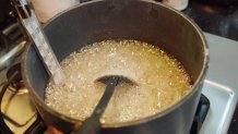 Frothy off-white liquid boils in a heavy metal pot with a spoon and a thermometer sticking out