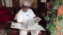 A woman in a white kitchen apron with glasses on her head sits in a chair and peruses a photo album