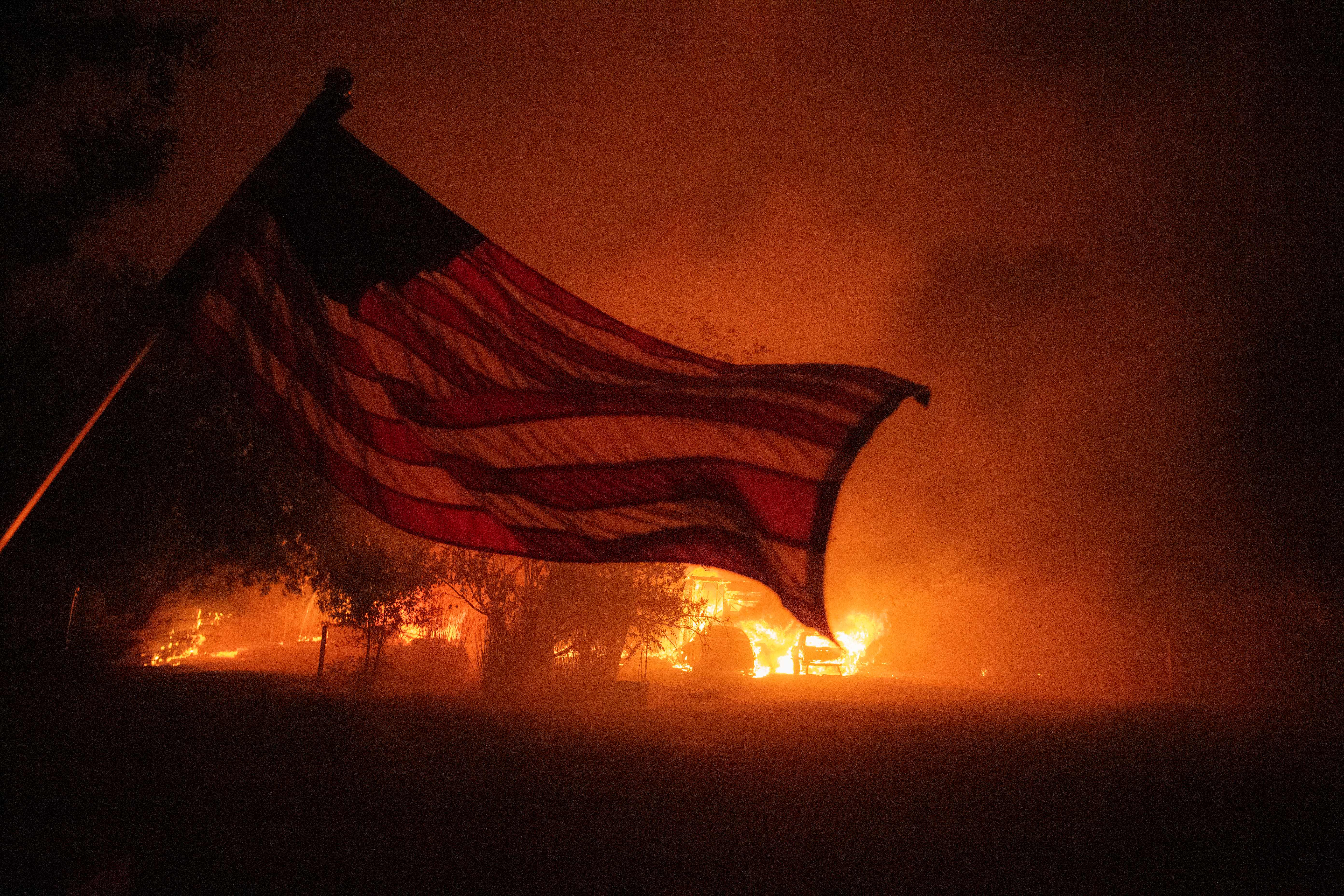 TOPSHOT – An American flag blows in the wind in front of a burning home in Vacaville, California during the LNU Lightning Complex fire on August 19, 2020. – As of the late hours of August 18,2020 the Hennessey fire has merged with at least 7 fires and is now called the LNU Lightning Complex fires. Dozens of fires are burning out of control throughout Northern California as fire resources are spread thin. (Photo by JOSH EDELSON / AFP) (Photo by JOSH EDELSON/AFP via Getty Images)