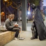 A man dressed as Batman engages in conversation with an older man seated on a low concrete wall next to a downtown building at night.