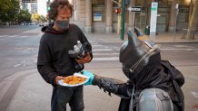 Kneeling man in a Batman helmet hands a plate of fried chicken and baked beans to a standing man in a black hooded sweatshirt