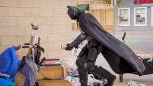 A man dressed as Batman kneels down to hand a can of spaghetti and meatballs to a man living in a small encampment against the side of a building