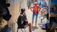 A person in a Spider-Man costume stands on the sidewalk, talking to a woman seated in a folding chair.