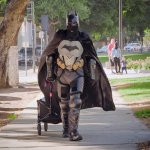 A man dressed as Batman pulls a small wagon through a park