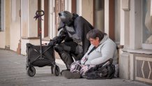 A man in a Batman costume kneels next to a woman sitting on the sidewalk against a building.