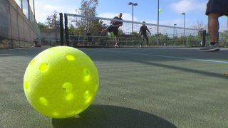 A yellow pickleball sits in the foreground as people play on a court behind it