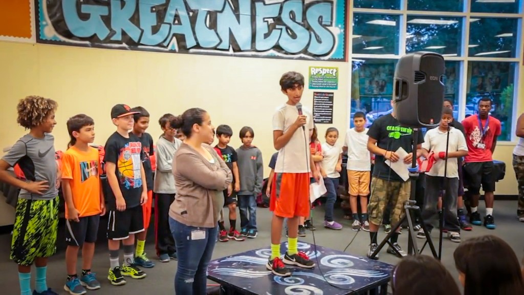 a 5th grade boy stands on a small stage in front of a gathering of other kids his age