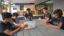 a group of middle school boys sits around a table outdoors with a laptop and a tablet