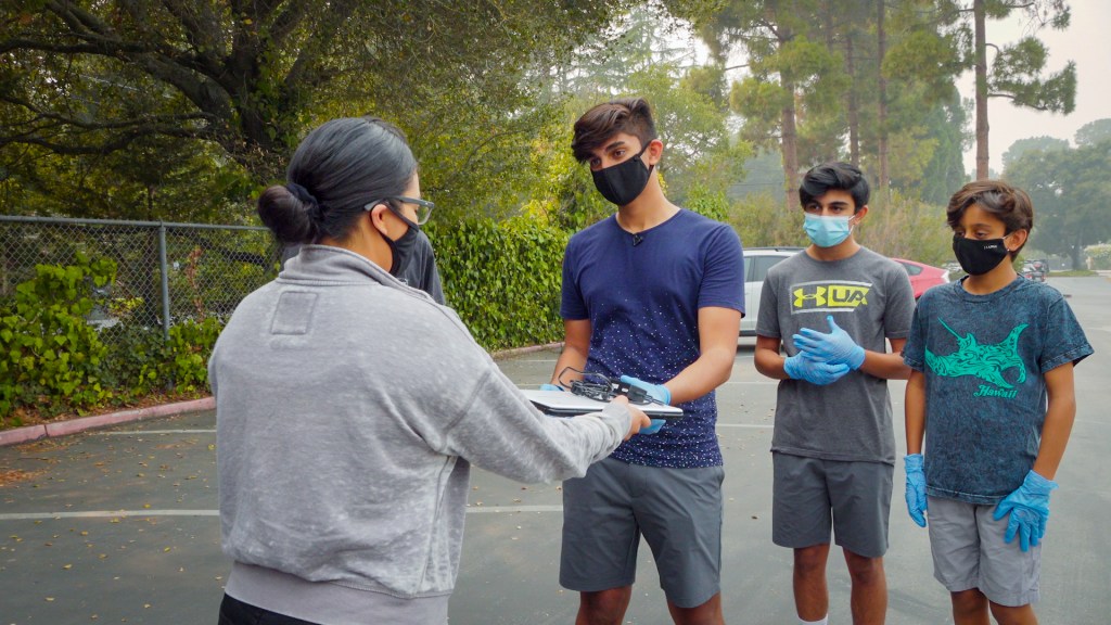 a group of teens hand a laptop computer to a woman in a parking lot