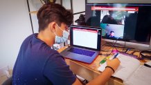 A teen sits at a computer, filling out a worksheet with a pen and talking to his teacher over video chat