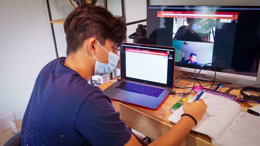 A teen sits at a computer, filling out a worksheet with a pen and talking to his teacher over video chat