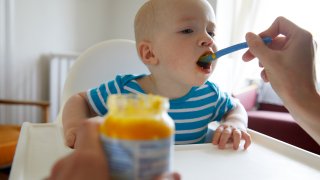 Baby taking a bite of food from a spoon with jar of baby food in foreground