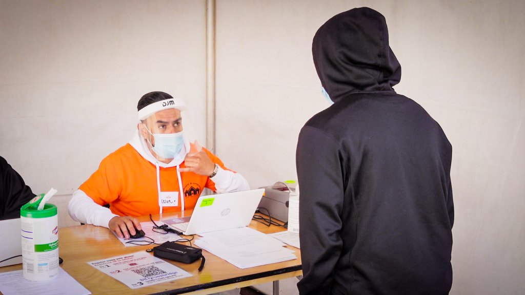 a man in an orange shirt, surgical mask and face shield sits at a desk as a person in a black hoodie approaches