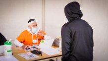 a man in an orange shirt, surgical mask and face shield sits at a desk as a person in a black hoodie approaches