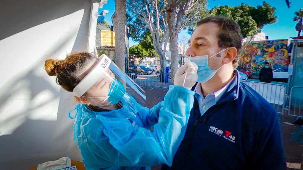 a woman in a face shield and disposable blue gown sticks a long swab up a reporter's nose