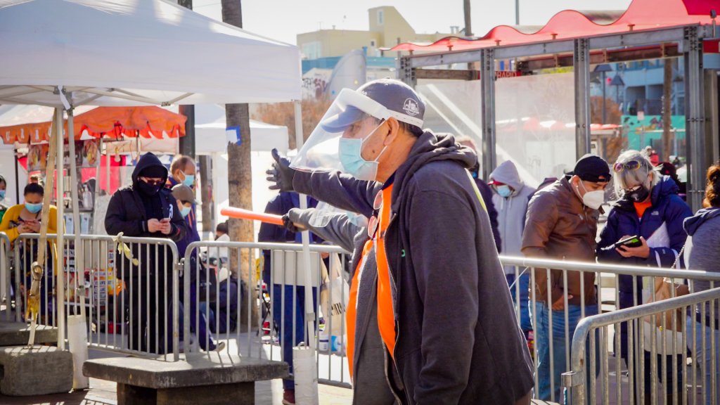 a man in an orange t-shirt and face shield directs the person at the front of the line past a line of barricades