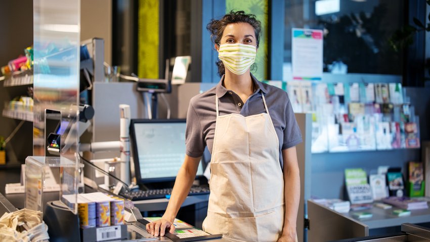 Portrait of female cashier wearing a protective face mask standing at checkout counter in a grocery store. Mature woman working at a supermarket during pandemic.