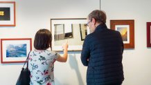 two people look at a photograph in a gallery that depicts a hand resting on the window sill of a house or apartment.