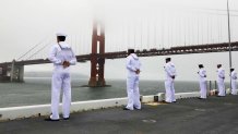 Sailors in white uniforms stand at the edge of the ship's deck as the ship sails under the Golden Gate Bridge