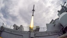 A missile launches from the central island of a Navy aircraft carrier.
