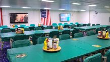 Tables and chairs with condiment racks covered in green cloth. An American flag and television monitors hang on the wall in the background.