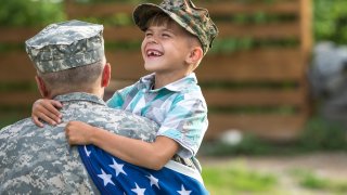 Military father hugs son holding American flag.