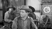 frame from a vintage western featuring a man in a hat sitting in a saloon, looking toward the entrance