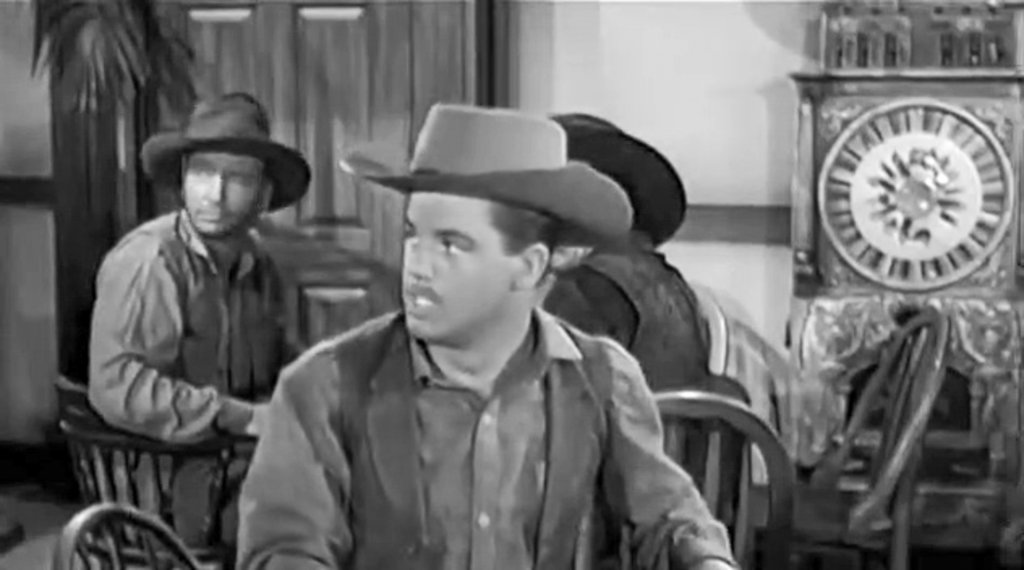 frame from a vintage western featuring a man in a hat sitting in a saloon, looking toward the entrance