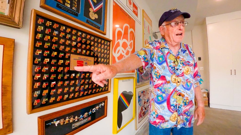 a man points to a wall of framed Olympic pins