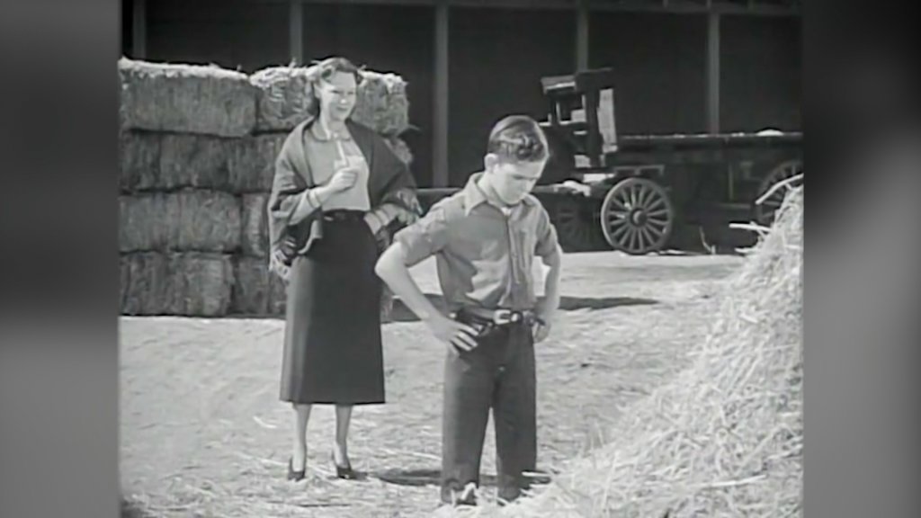 black and white frame of a young boy standing with his hands on his hips, staring at a haystack as his mother looks on from behind him