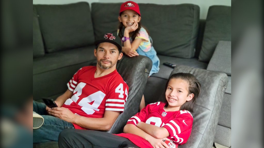 a father and his two kids sit in reclining chairs wearing 49ers jerseys.