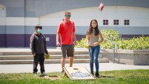 two kids and their teacher stand outside with a cornhole board between them while a bean bag drops from the sky