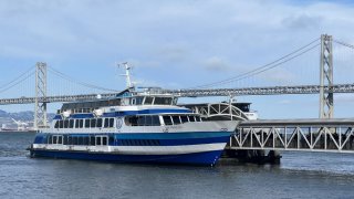 Blue and white ferry boat docked at the Port of San Francisco, with the Bay Bridge in the background, San Francisco, California, August 17, 2023.