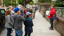 A group of 16 people stands around a tour guide in a red jacket on a sidewalk.