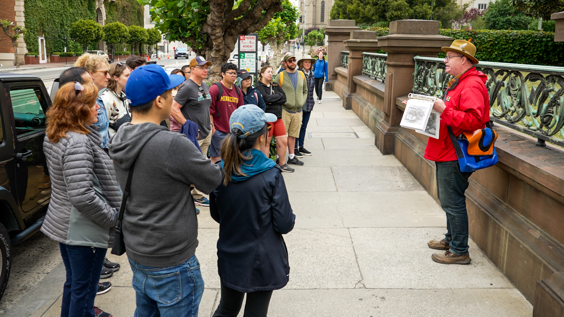 A group of 16 people stands around a tour guide in a red jacket on a sidewalk.