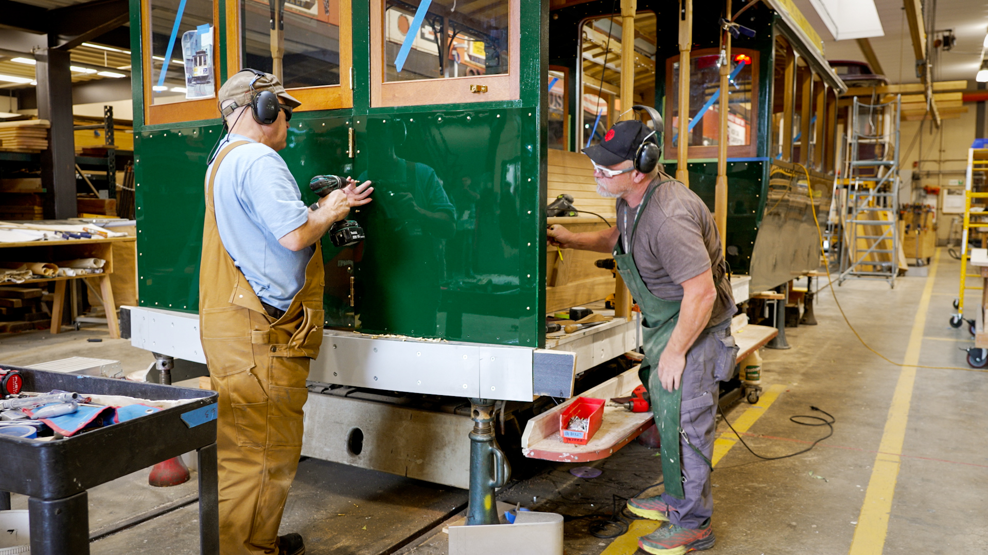 Two carpenters work on the front of a green half-built San Francisco cable car inside a workshop.
