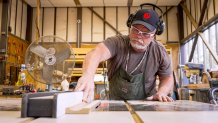 A carpenter with a white beard wearing safety glasses and protective earmuffs guides a long piece of wood across a table saw.