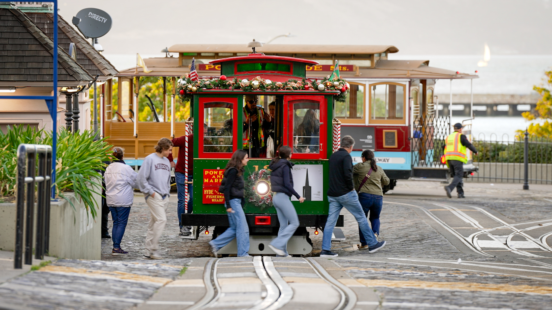 Passengers in casual dress walk around the front of a green and red cable car to board it. Behind them, another cable car is turned around on a turntable, and behind that, boats sail on the open waters of the Bay.