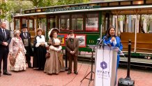 Four actors in Victorian costume stand in front of a green and white San Francisco cable car. A woman in a blue suit speaks at a lectern next to them.